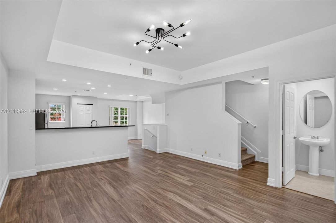 Open-concept interior featuring wood-finish flooring, a multi-arm modern chandelier, tray ceilings, and a powder room with a pedestal sink and circular mirror