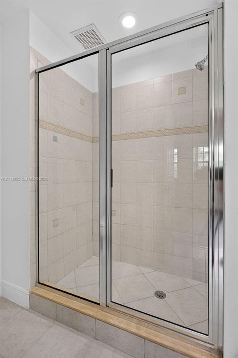 Walk-in shower enclosure featuring a polished metal frame, light-toned ceramic wall tiling with a decorative border inlay, diamond-patterned floor tile, and a raised stone threshold