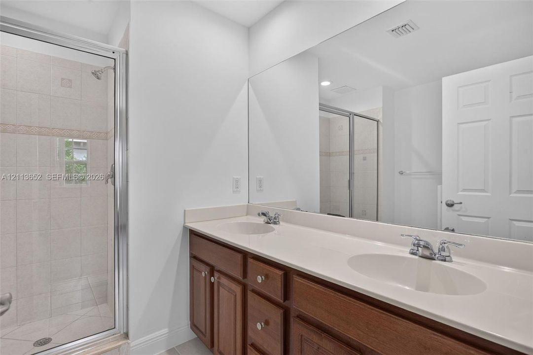 Double vanity with integrated sinks, neutral countertop, wood cabinetry, and a full-width wall mirror, alongside a walk-in shower with tiled surround and glass enclosure