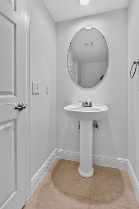 White pedestal sink featuring a chrome dual-handle faucet, oval wall-mounted mirror, and tile flooring