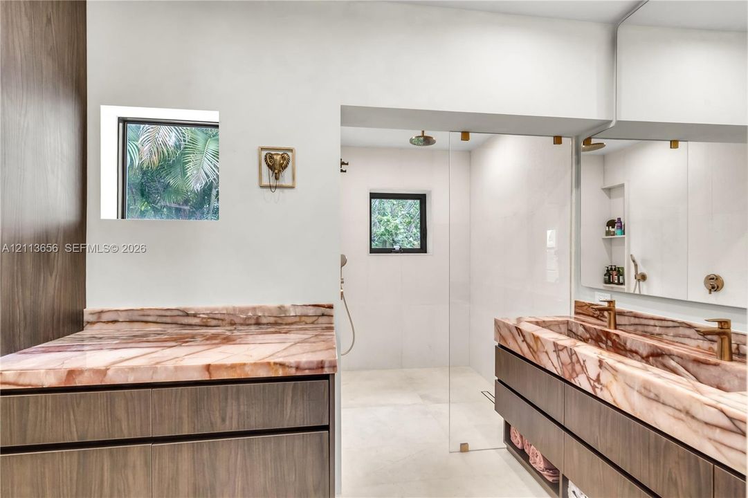 Floating vanity with integrated stone-finish dual sinks, stone countertops featuring distinct veining, flat-panel wood-finish cabinetry, a large frameless mirror, and a walk-in shower with a glass enclosure