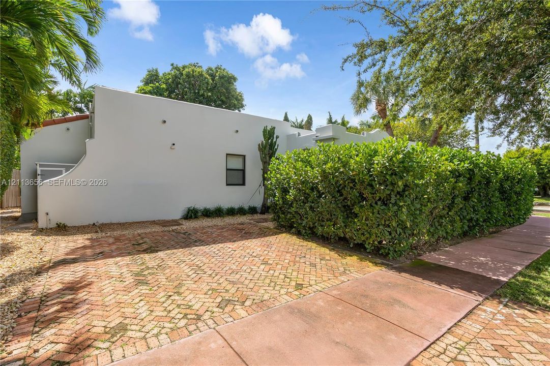 Stucco exterior featuring a parapet roofline, herringbone-patterned brick driveway, and mature privacy hedge