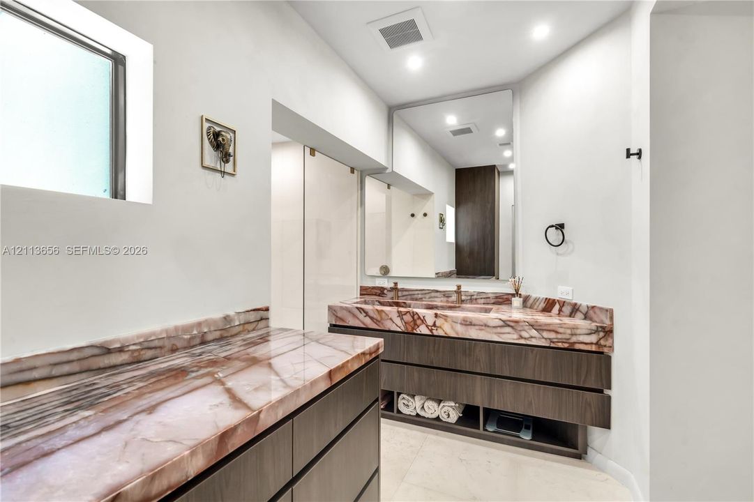 Dual-sink vanity featuring dark wood-finish cabinetry, extensive stone-patterned countertops, a large frameless mirror, and recessed ceiling lighting