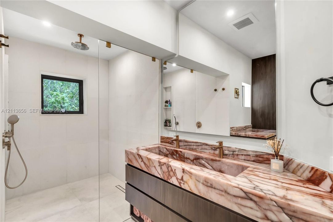 Floating vanity with integrated stone-finish double sink, floor-to-ceiling glass-enclosed shower, wall-mounted faucet fixtures, built-in recessed wall shelving, and expansive wall-to-wall mirror