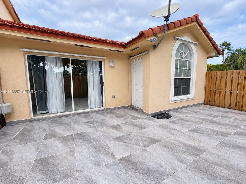 Stucco exterior featuring red clay roof tiles, large-format grey tile patio, sliding glass door, arched window, and wood privacy fencing