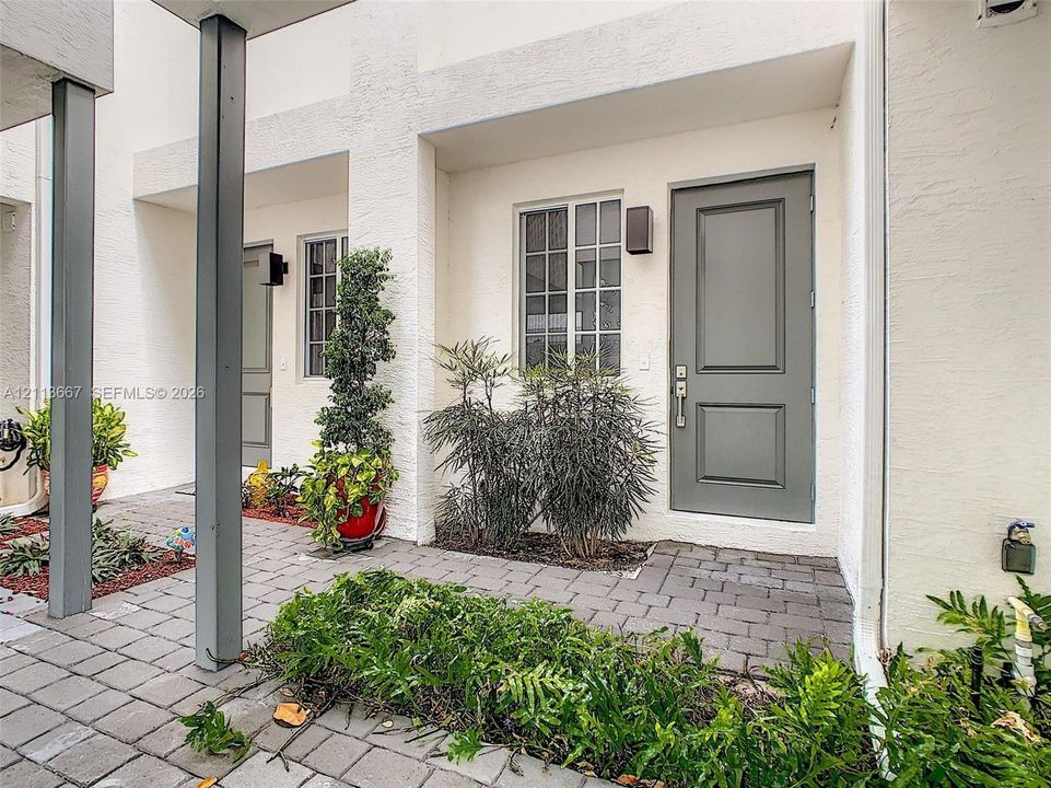 Exterior entry featuring a textured stucco facade, paver walkway, multi-pane windows, and a solid-panel door with architectural sconce lighting