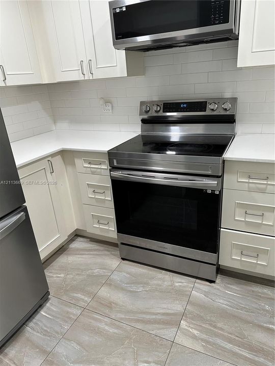 Kitchen featuring white shaker-style cabinetry, solid-surface countertops, a subway tile backsplash, stainless steel appliances, and large-format porcelain tile flooring