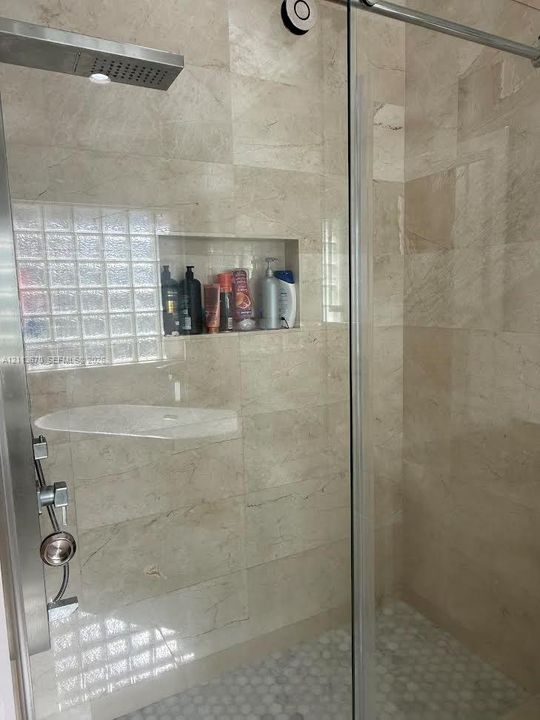 Walk-in shower featuring natural stone wall tile, built-in recessed wall niche, rainfall shower head, glass-block window, and pebble-style floor tile