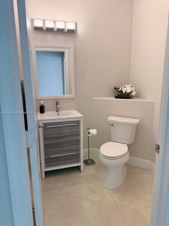 Vanity featuring a textured grey cabinet base, rectangular basin, and single-handle faucet, paired with a white-framed mirror, four-light vanity fixture, and neutral stone-finish tile flooring