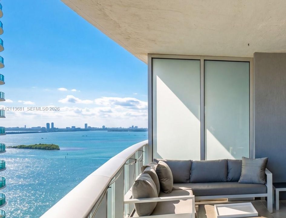 Covered balcony featuring a glass panel railing, concrete ceiling, and floor-to-ceiling glass sliding doors, with panoramic water and skyline scenery