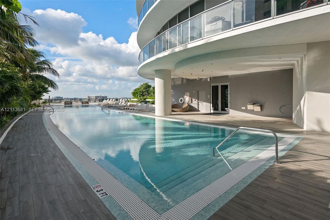 Curved swimming pool featuring a tile border, metal handrails, integrated steps, and wood-finish decking