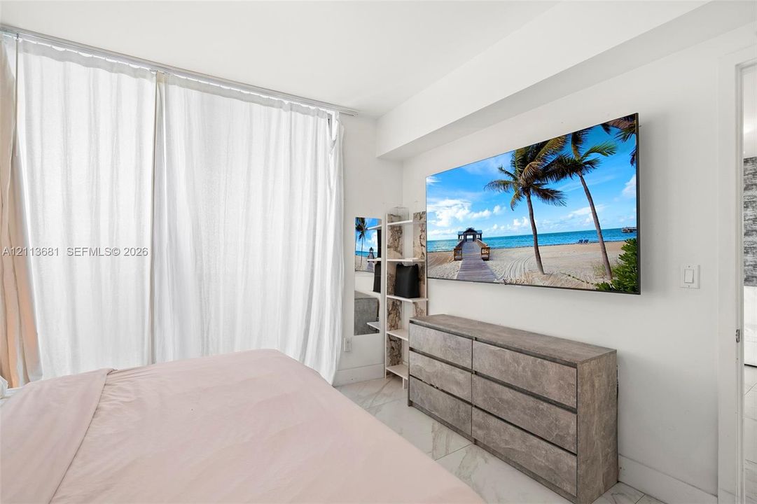 Floor-to-ceiling windows featuring sheer curtains, polished tile flooring, and wall-mounted television