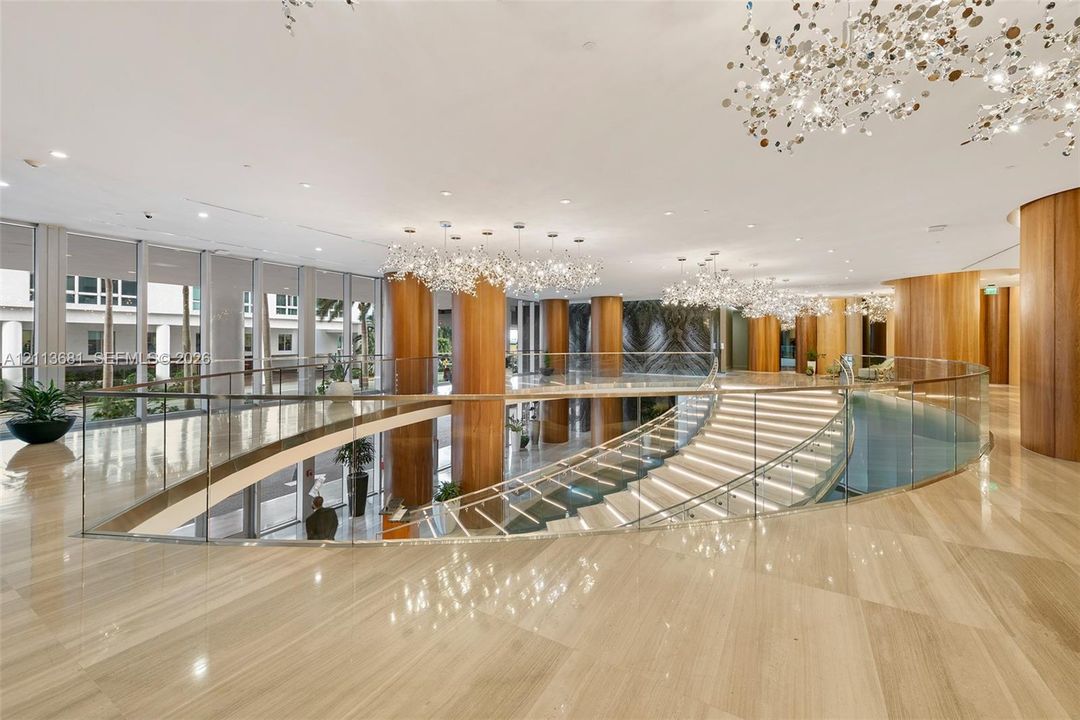 Grand atrium featuring polished stone flooring, custom architectural wood-finish columns, a sweeping curved staircase with glass railing, and oversized decorative chandeliers