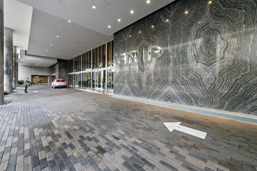 Covered motor court entrance featuring natural stone wall cladding, floor-to-ceiling glass paneling, overhead recessed lighting, and paver brick driveway