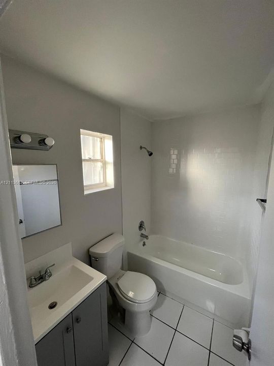 Bathroom featuring a grey vanity with an integrated sink, white tile flooring, white wall-surround bathtub, and dual-light vanity fixture