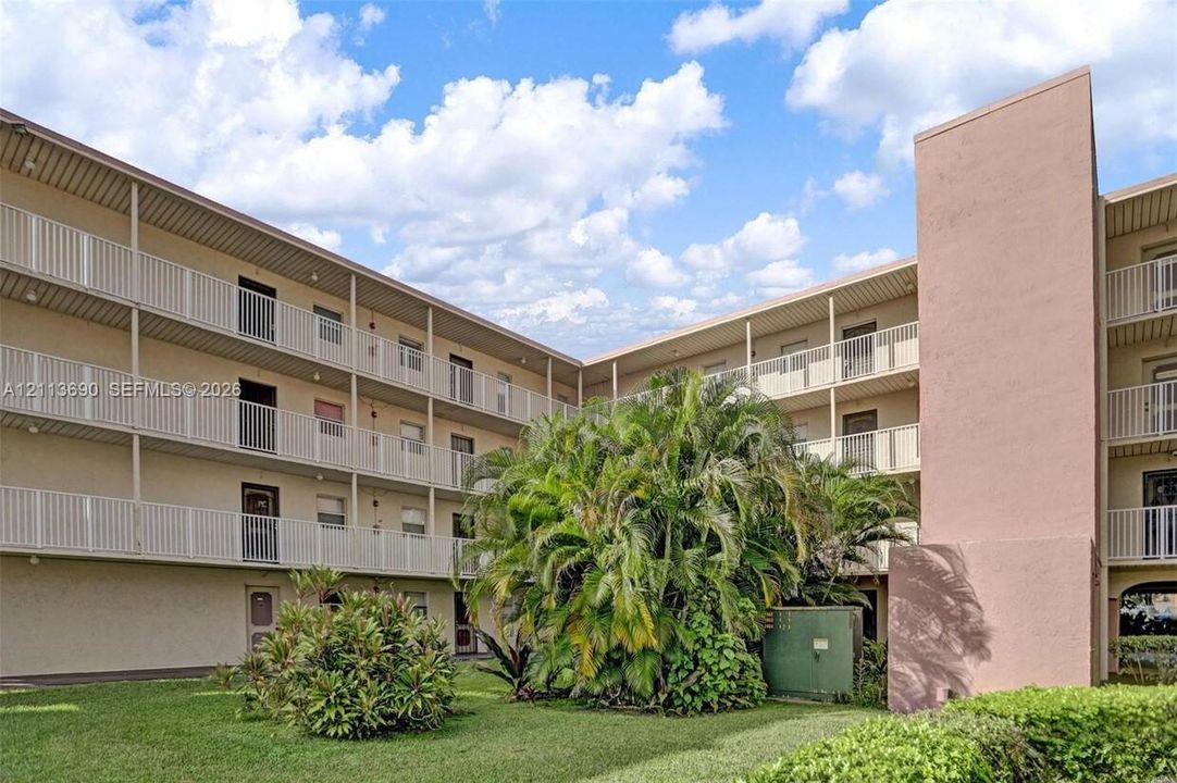 Multi-story building exterior featuring a stucco facade, tiered white metal railings, and integrated balcony levels, surrounded by mature palms and landscaping