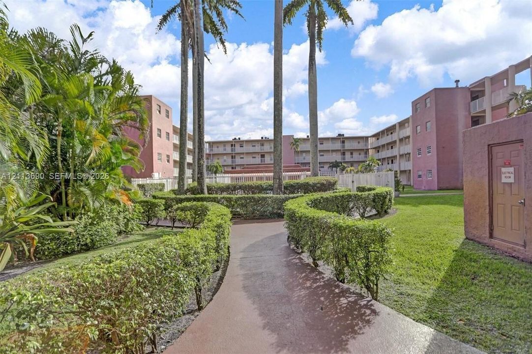 Paved walkway lined with manicured hedges, mature palm trees, and landscaped grounds