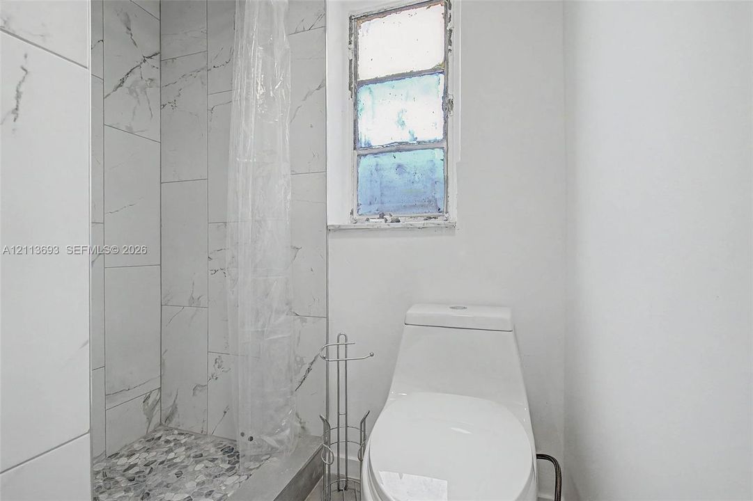 Walk-in shower featuring large-format stone-patterned tile, pebble-tiled flooring, and a single-piece elongated toilet