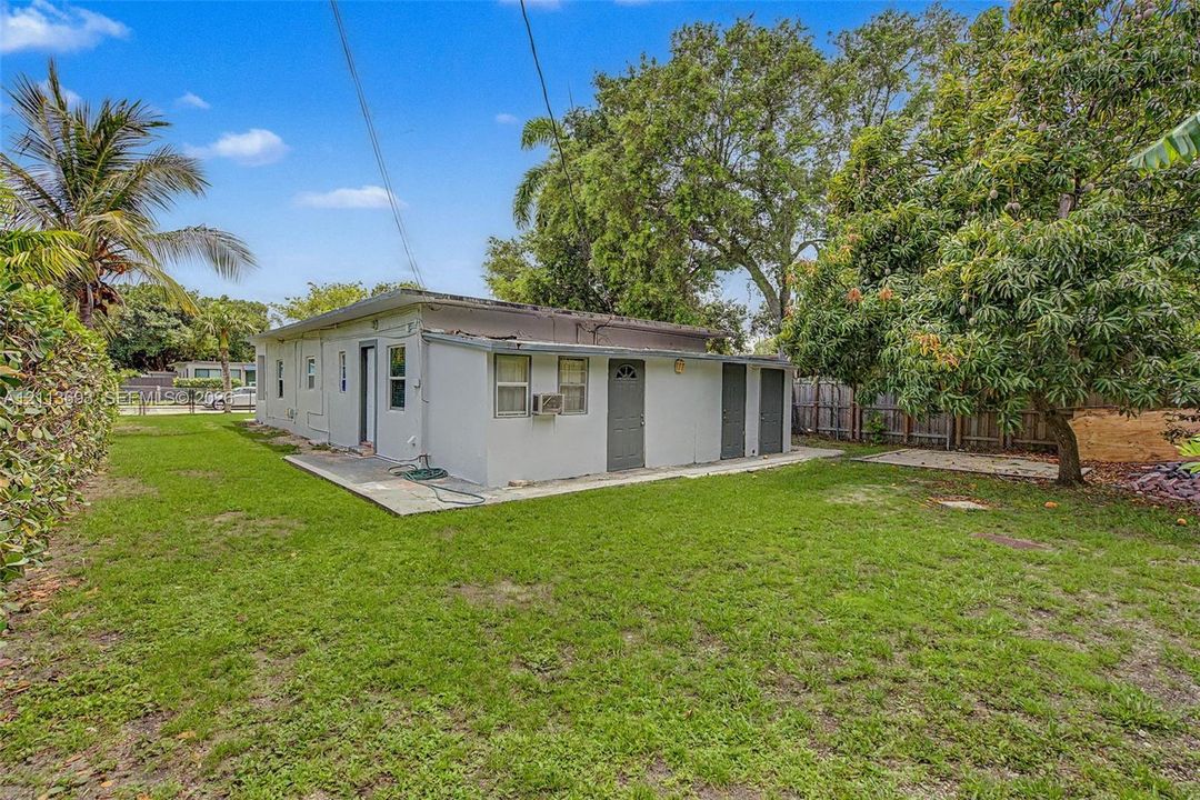 Rear exterior featuring a concrete patio, exterior access doors, mature trees, and a perimeter fence