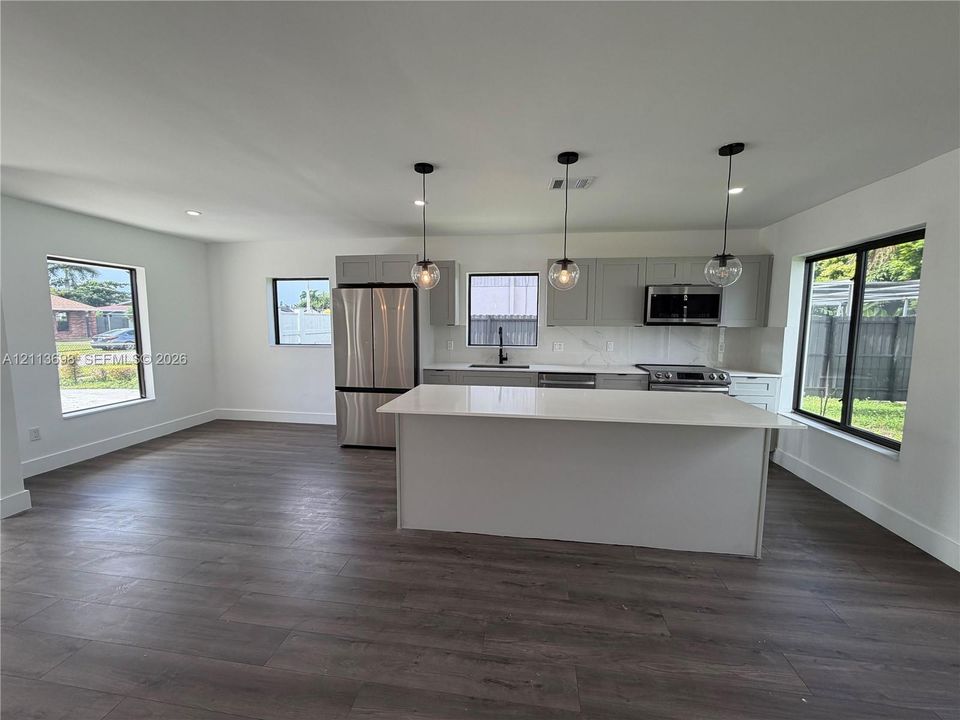 Kitchen featuring wood-finish flooring, quartz-surfaced island, shaker-style cabinetry, stainless steel appliances, and glass pendant lighting