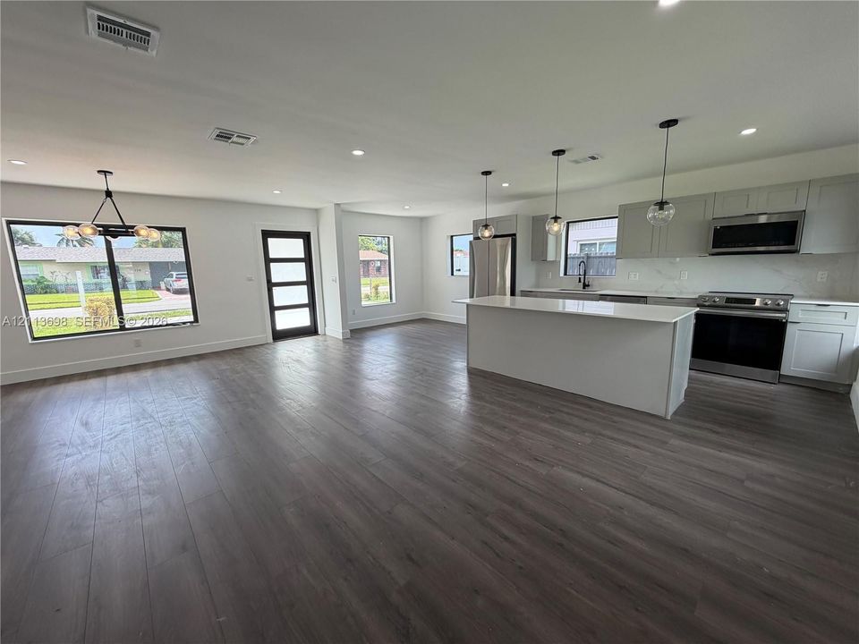 Open floor plan featuring wood-finish flooring, a kitchen island with stone-surface countertop, flat-panel cabinetry, stainless steel appliances, and multi-pendant lighting
