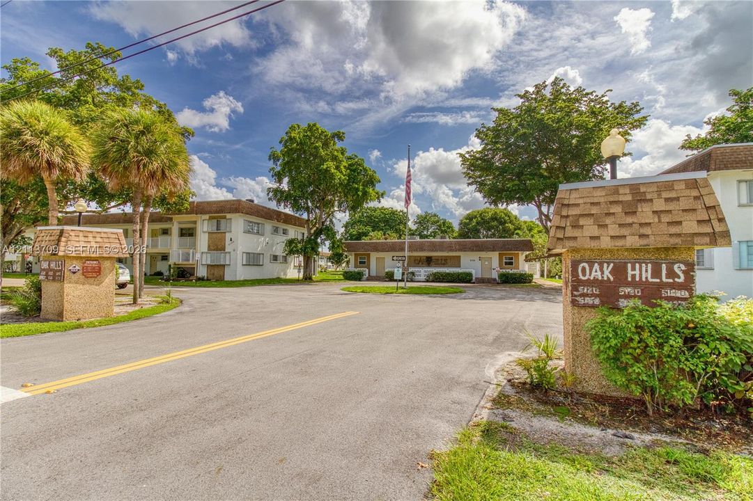 Multi-building property entrance featuring textured masonry signage, mature palm trees, asphalt paving, and established landscaping