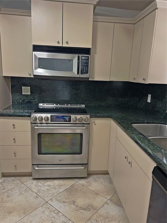 Kitchen featuring stone countertops, stainless steel appliances, upper and lower cabinetry, a dual-basin undermount sink, and tile flooring