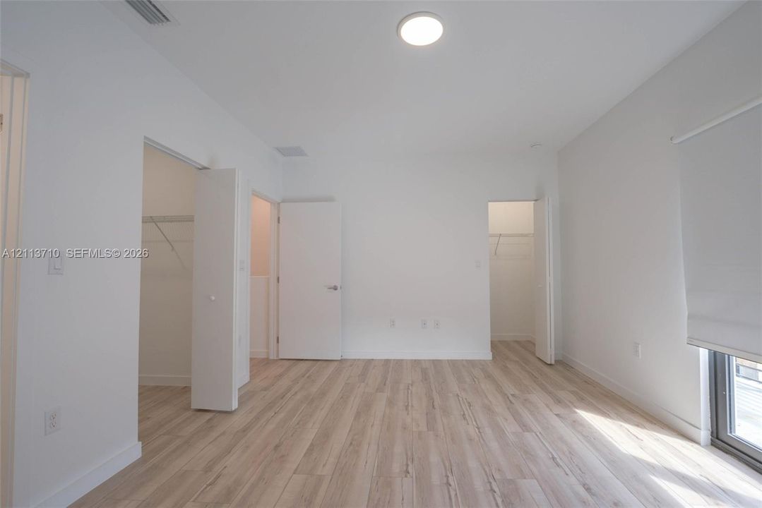 Room featuring wood-finish flooring, two walk-in closets with built-in shelving, flush-mount ceiling lighting, and a window fitted with a roller shade