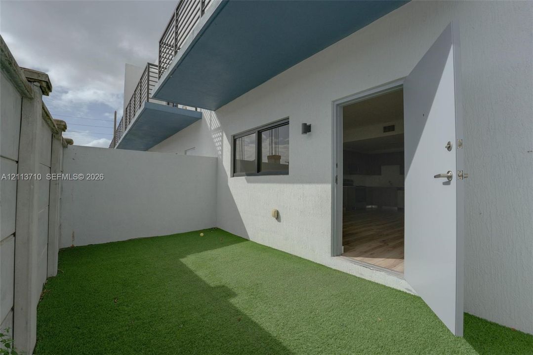 Private courtyard featuring artificial turf, white stucco exterior walls, a metal privacy fence, and a cantilevered upper-level balcony with metal railing
