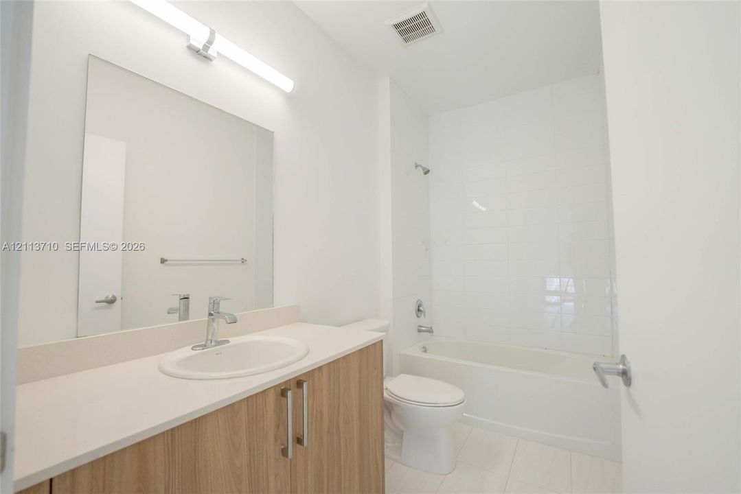 Bathroom featuring a vanity with a light-toned countertop, oval undermount sink, wood-finish cabinetry, a full-width mirror with linear lighting, and a bathtub with tile surround
