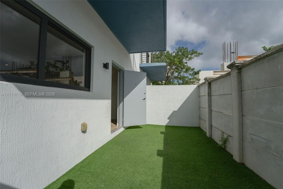 Exterior area featuring synthetic turf, smooth white stucco walls, dark-framed windows, and a precast concrete privacy wall