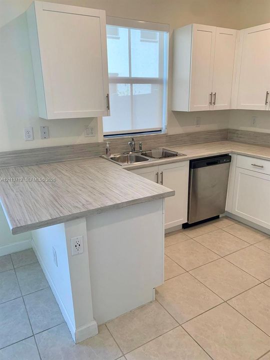 Kitchen featuring white shaker-style cabinetry, neutral-toned countertops with matching backsplash, stainless steel dishwasher, dual-basin stainless steel sink, and light-colored tile flooring