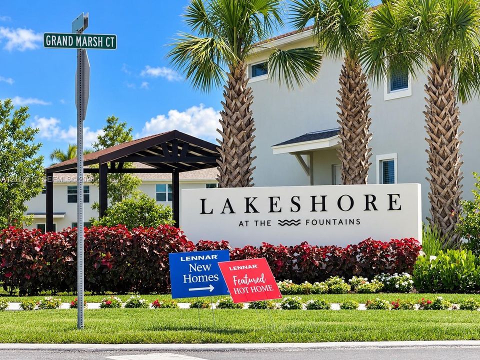 Community monument signage featuring a light-colored stucco finish, manicured landscaping with mature palm trees, and structural shaded entry canopy