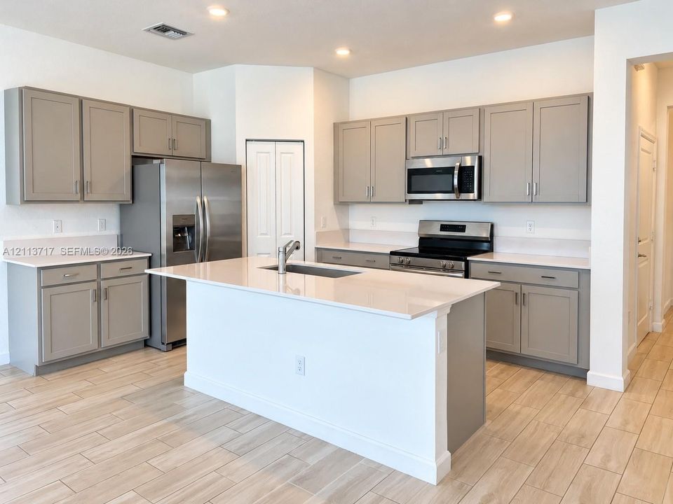 Kitchen featuring grey shaker-style cabinetry, quartz countertops, a central island with an undermount sink, stainless steel appliances, and wood-finish tile flooring