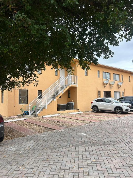 Exterior featuring a stucco facade, external staircase with metal railings, and a brick-patterned paved parking area with designated spaces