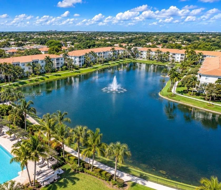 Aerial perspective featuring a central lake with a decorative fountain, surrounding multi-story buildings with tile roofing, perimeter walkways, and mature palm landscaping