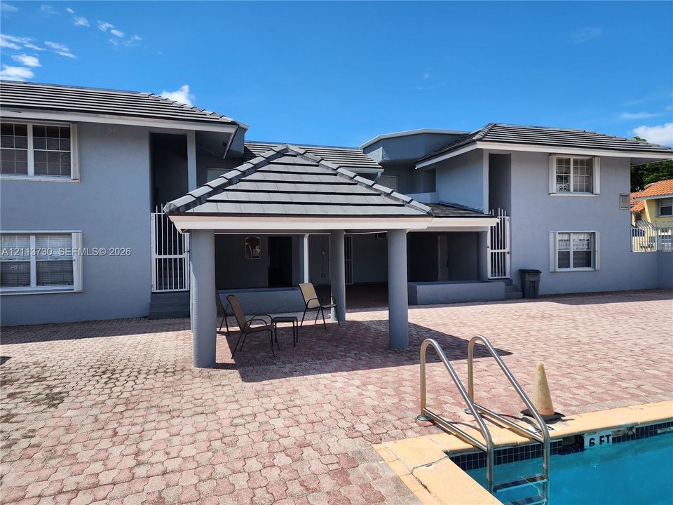 Stucco exterior with multi-pitched tile roofing, covered poolside pavilion with concrete columns, and expansive stone paver patio