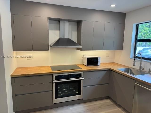 Modern kitchen featuring flat-panel cabinetry, wood-finish countertops, stainless steel wall oven, matching range hood, and a stainless steel undermount sink with a gooseneck faucet