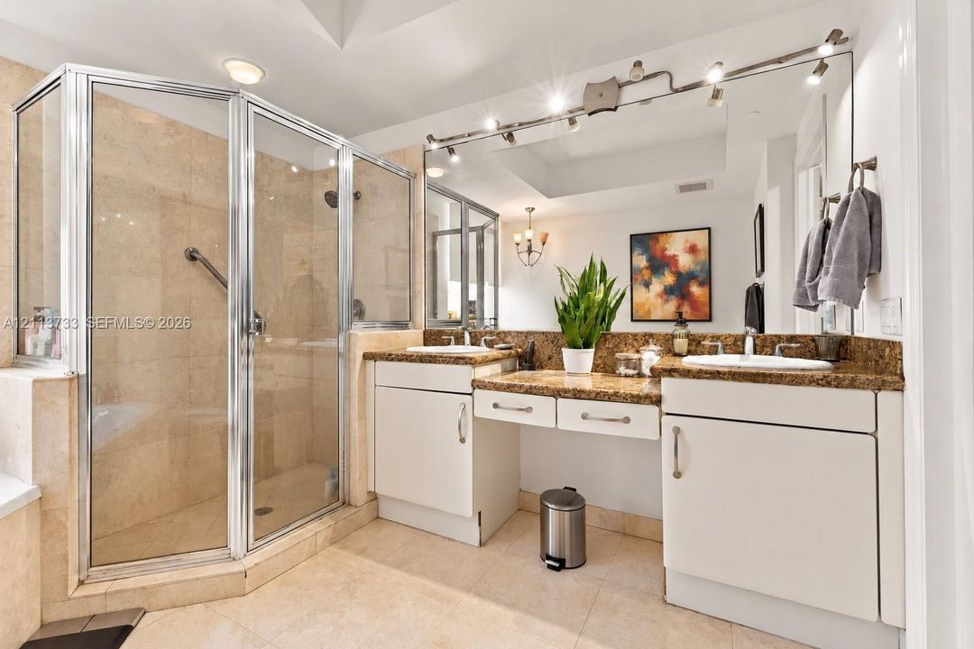 Double vanity featuring stone countertops with dual undermount sinks, central dressing table, full-width mirror with integrated track lighting, and a walk-in glass-enclosed shower with tile surround