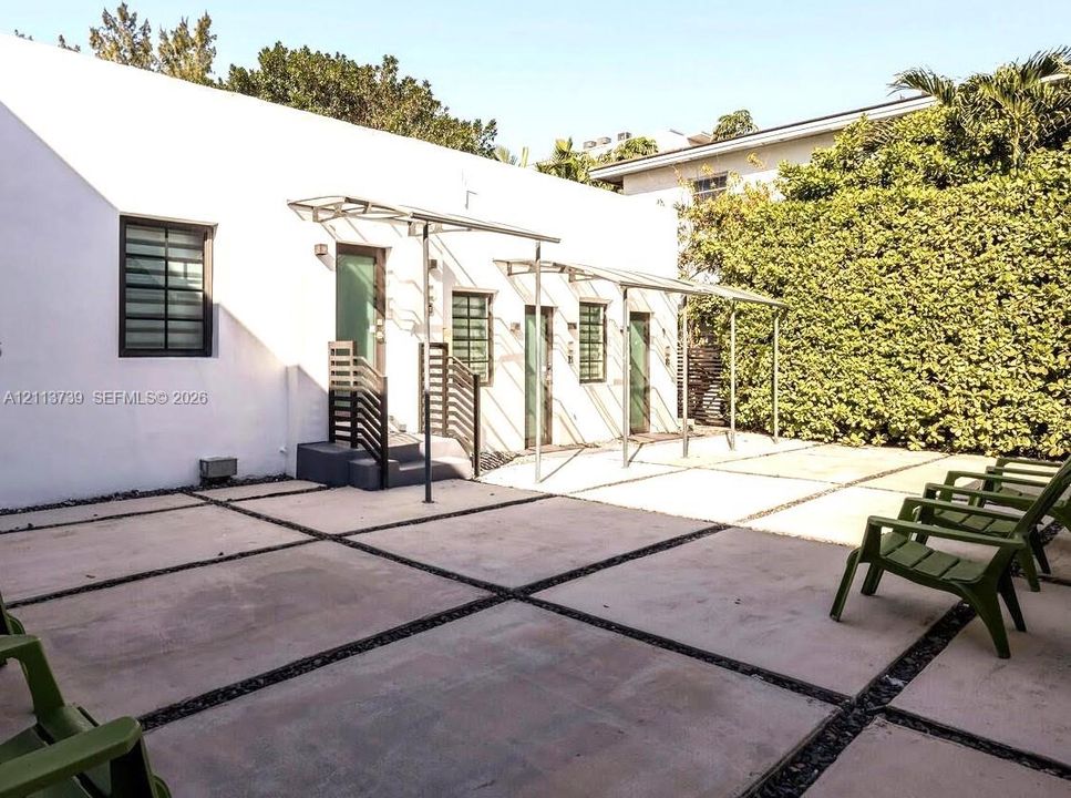 Exterior courtyard featuring large-format concrete pavers with gravel inlay, metal-framed awnings, three individual entryways with steps and railings, and perimeter hedge screening