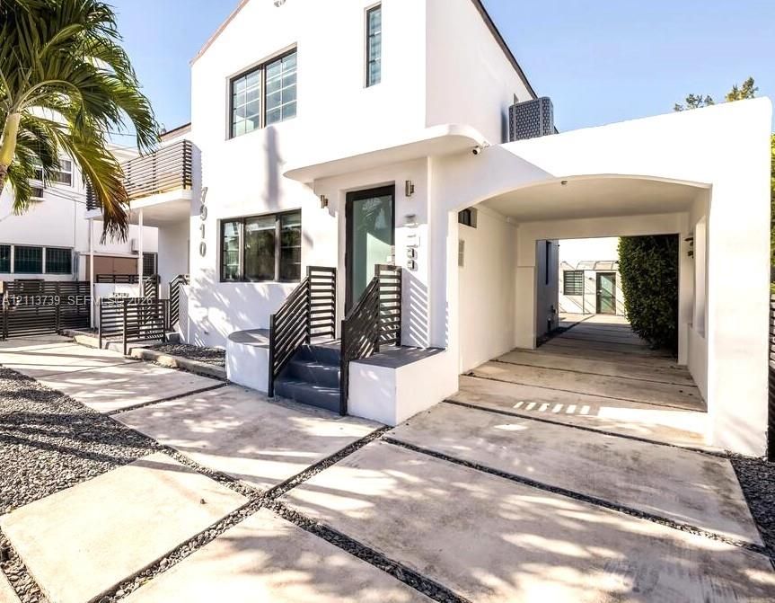 Modern exterior featuring a smooth stucco facade, arched covered parking, private balcony with horizontal slat railings, and a concrete paver driveway with decorative stone accents