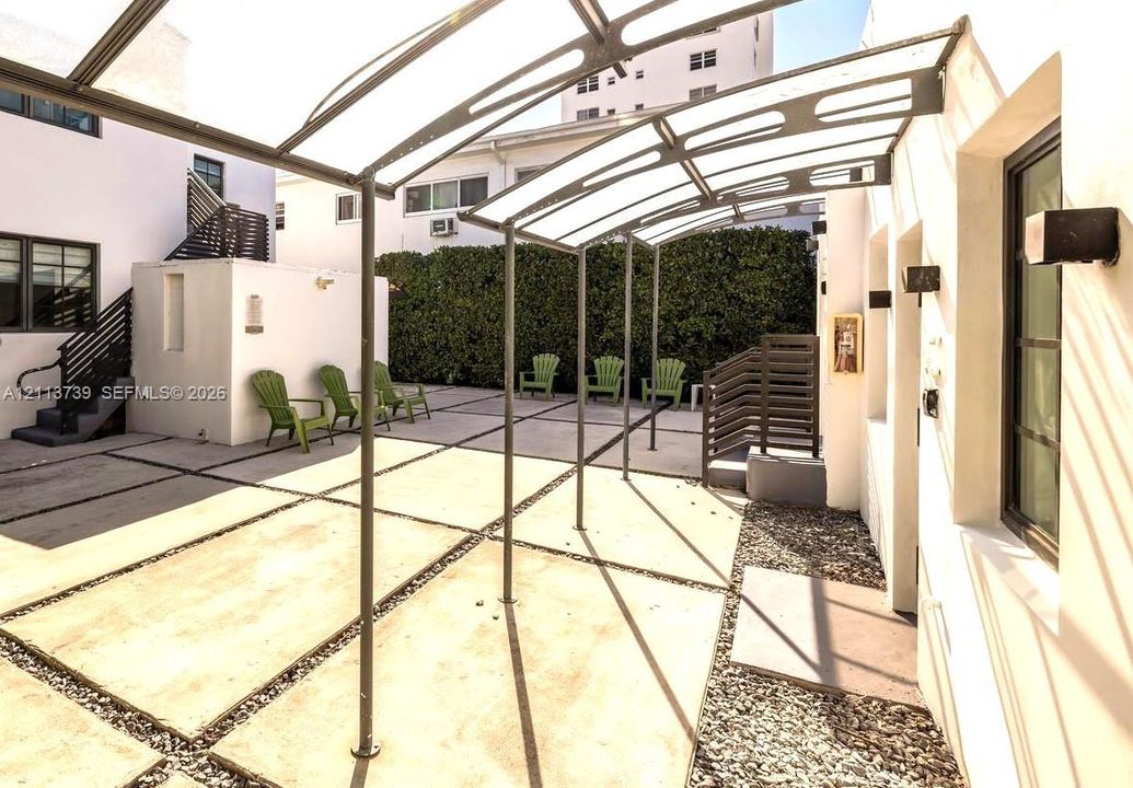 Outdoor courtyard featuring large-format concrete pavers, a metal-frame structural canopy, exterior wall-mounted lighting, and a perimeter landscape hedge