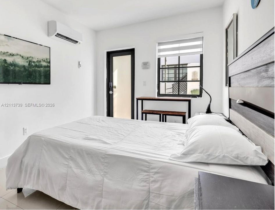 Wall-mounted ductless air conditioning unit, black-trimmed glass-panel exterior door, multi-pane window with integrated roller shade, and tile flooring