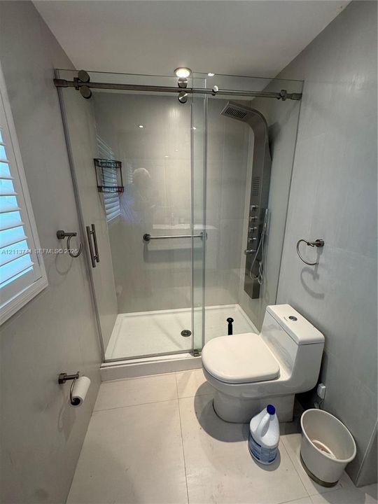 Walk-in shower with a sliding glass door, a wall-mounted stainless steel multi-head shower panel, integrated grab bar, large-format tile flooring, and a dual-flush toilet