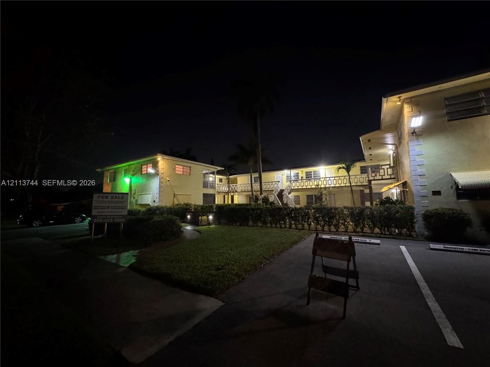Multi-story exterior featuring stucco siding, metal railing balconies, paved parking area, and mature palm trees
