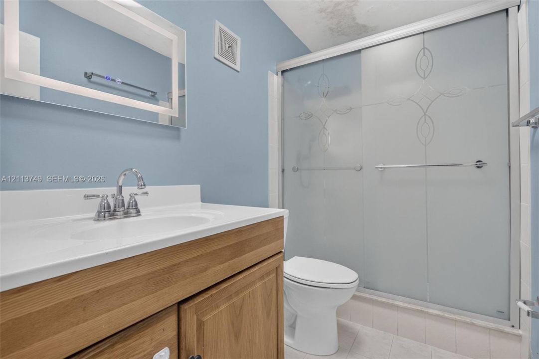 Wood-finish cabinetry, solid-surface integrated countertop and sink, brushed-metal faucet, wall-mounted backlit mirror, and glass shower enclosure with decorative etching