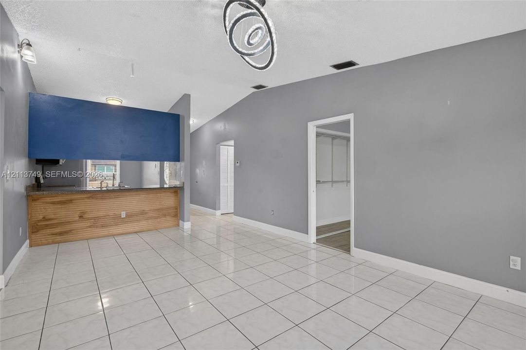 Vaulted ceiling featuring a multi-ring LED chandelier, neutral ceramic tile flooring, and a kitchen pass-through counter with a wood-finish facade