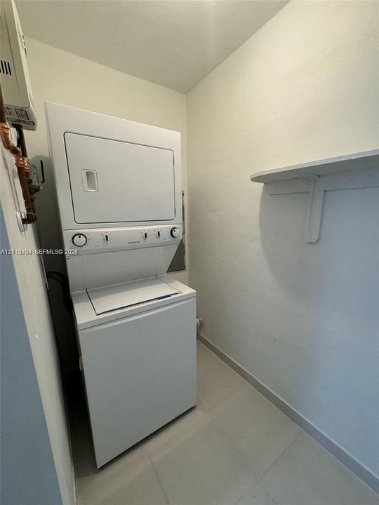 Laundry area featuring a white stackable washer and dryer unit, neutral tile flooring, and a wall-mounted storage shelf