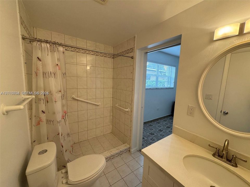 Bathroom featuring a tile-surround walk-in shower with safety grab bars, vanity with an integrated sink and neutral countertop, wall-mounted circular mirror, and overhead vanity lighting