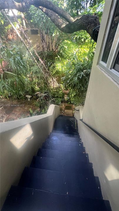 Exterior staircase with dark-finished treads, solid side walls, and a mounted metal handrail leading to a secluded garden area with mature trees, stone paving, and lush vegetation